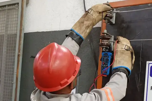 Profissional com capacete e luvas isolantes realizando medição em painel elétrico
