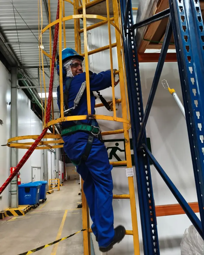 Participante de treinamento de NR-35 subindo escada marinheiro com cinto paraquedista e talabarte duplo, em armazém de empresa cliente